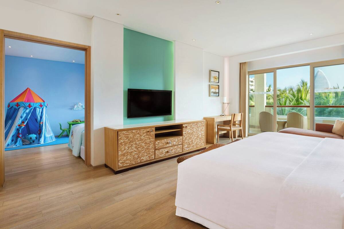 Een moderne hotelkamer met een groot bed, een tv bevestigd aan een houten kast, een bureau met een stoel, en uitzicht op palmbomen door grote ramen.