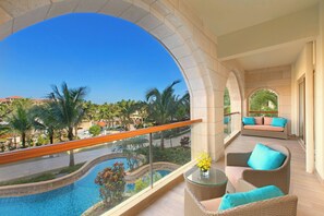 Ein Balkon mit freiem Blick auf einen Swimmingpool, ein Korbssessel mit Kissen und ein Glastisch mit Blumen in einer Vase.
