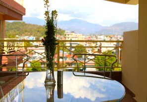 A balcony with a glass table and chairs, a vase with flowers, and a view of a cityscape with mountains in the background. - Hotel Splendid View (Pokhara)