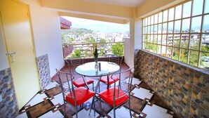 A balcony with a round table and red chairs, a view of a cityscape, and a tiled floor.