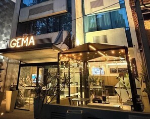 A modern building with a 'GEMA' sign, a glass entrance, and outdoor seating with plants. - Gema Luxury Suites (Montevideo)