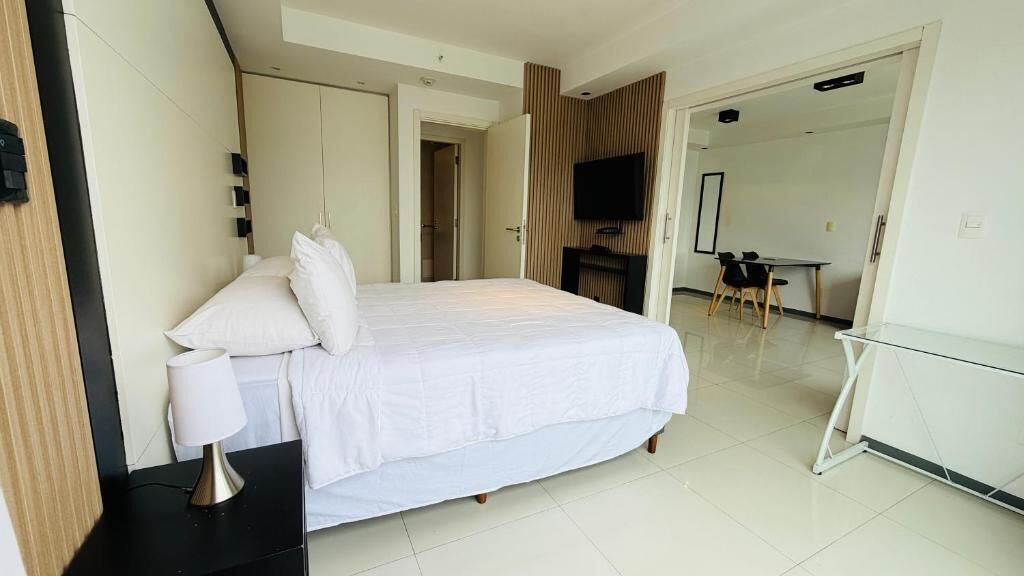 A hotel room with a large bed, a bedside table with a lamp, a TV mounted on the wall, and a desk with chairs.