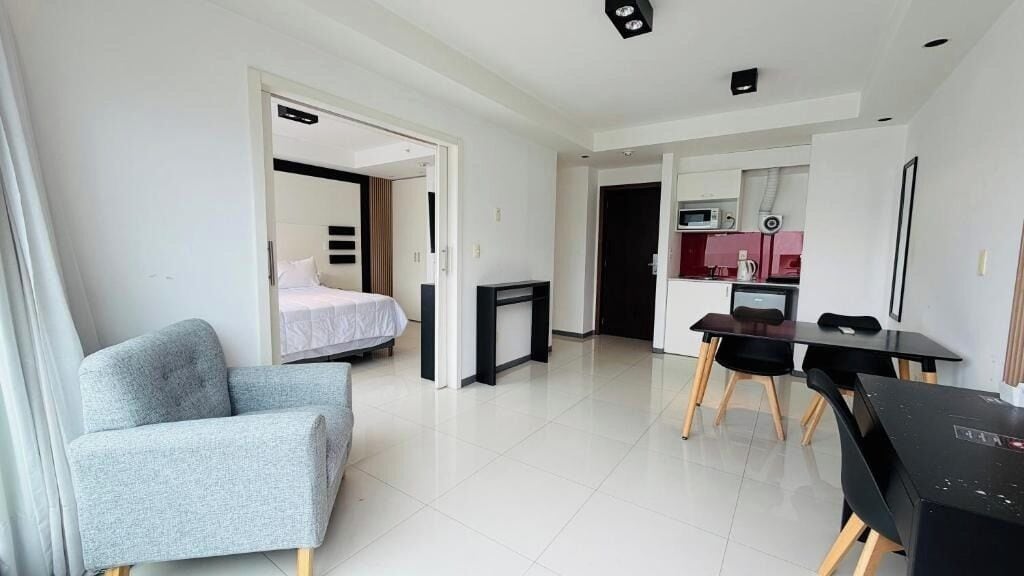 A modern hotel room with a bed, a grey armchair, a dining table with chairs, and a kitchenette area.