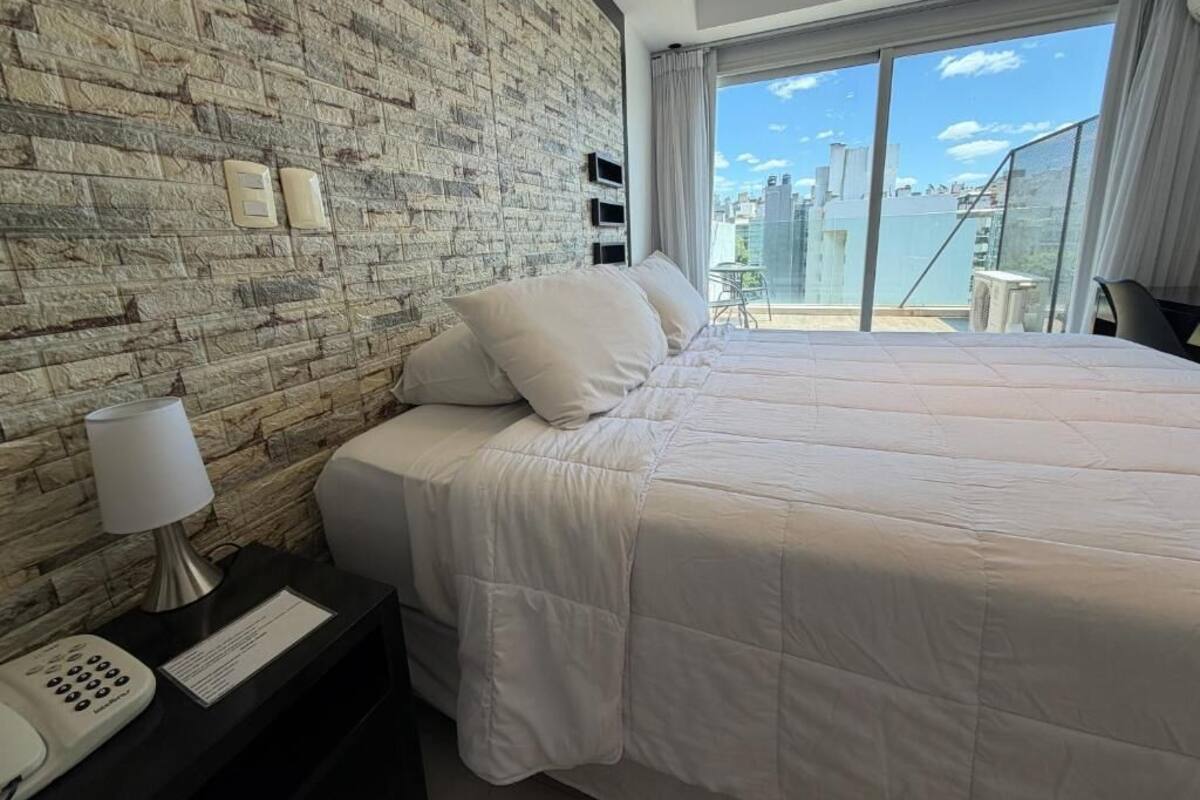A hotel room with a large bed, a stone wall, a balcony with a city view, and a telephone on the nightstand.