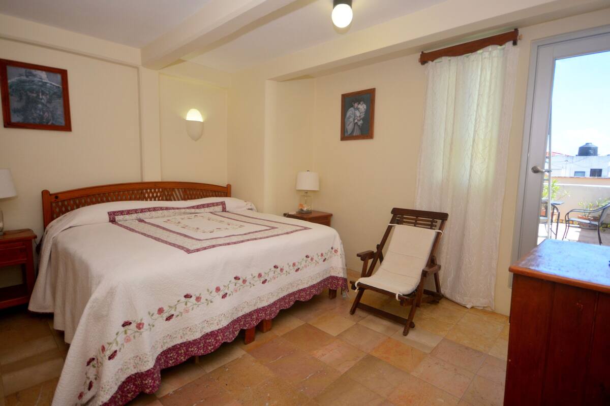 A bedroom with a bed, a chair, a nightstand, a lamp, and a door leading to a balcony.