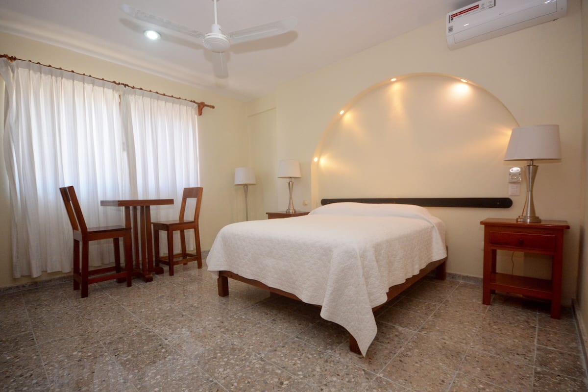 A bedroom with a bed, a desk, two chairs, a nightstand, and a ceiling fan.