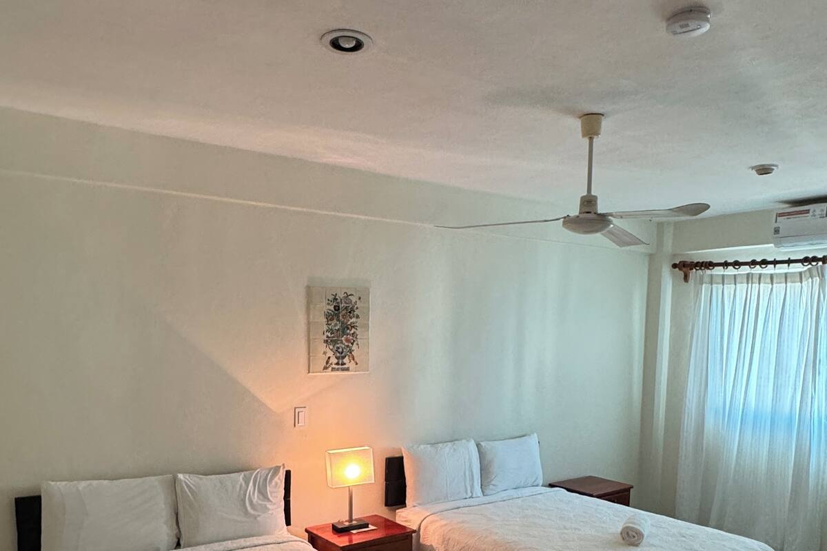 A hotel room with two beds, a ceiling fan, a nightstand with a lamp, and a window with curtains.