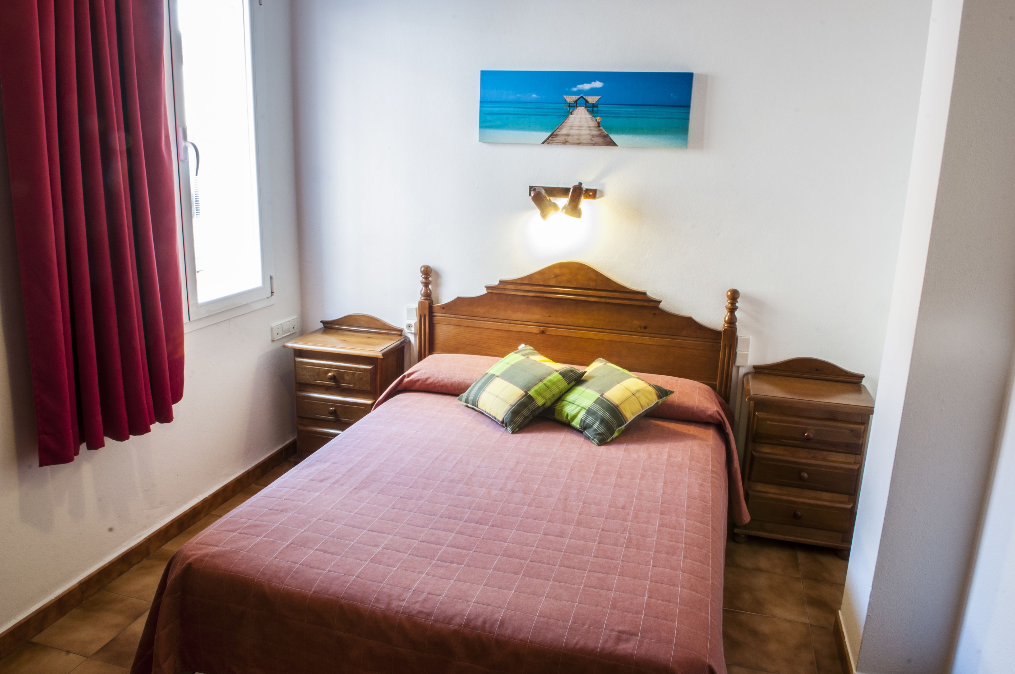 A bedroom with a wooden bed, two nightstands, a wall-mounted lamp, a window with curtains, and a painting of a pier.