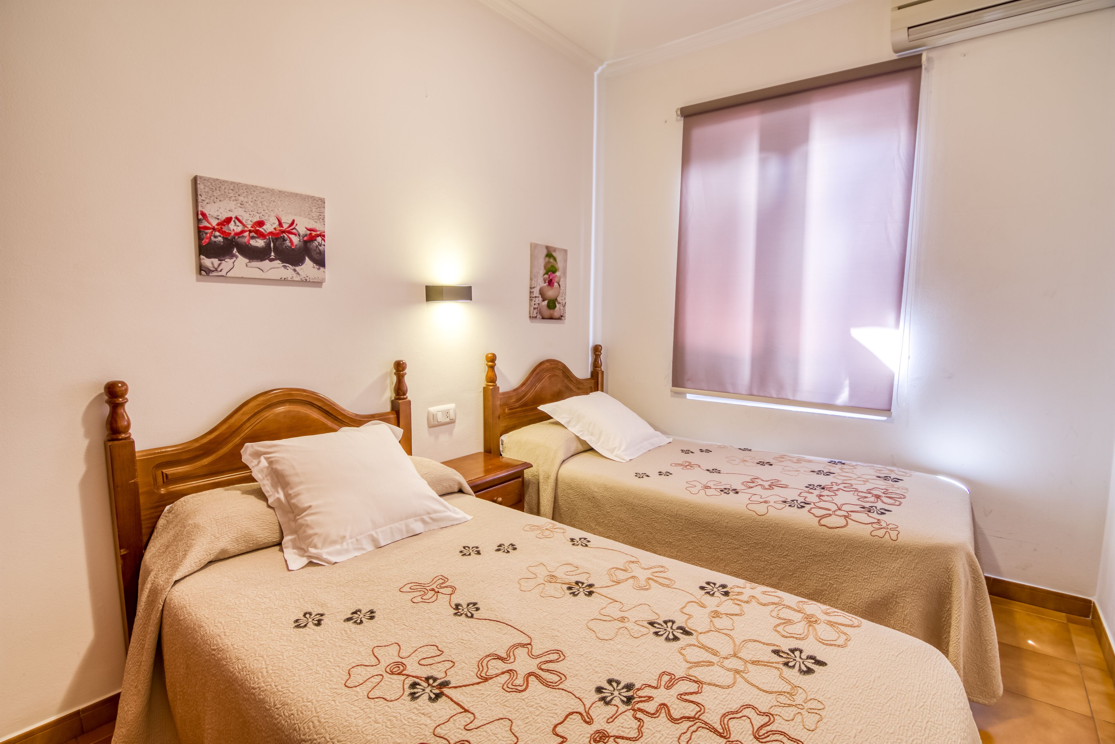 Una habitación con dos camas de madera, cada una con una colcha estampada y almohadas blancas. Hay una lámpara de pared y una ventana con cortina.