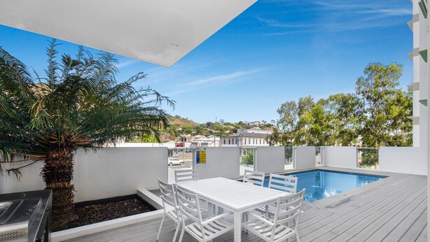 Un espace piscine aménagé sur le toit, avec une table et des chaises blanches, entouré d’une terrasse en bois et d’un palmier.
