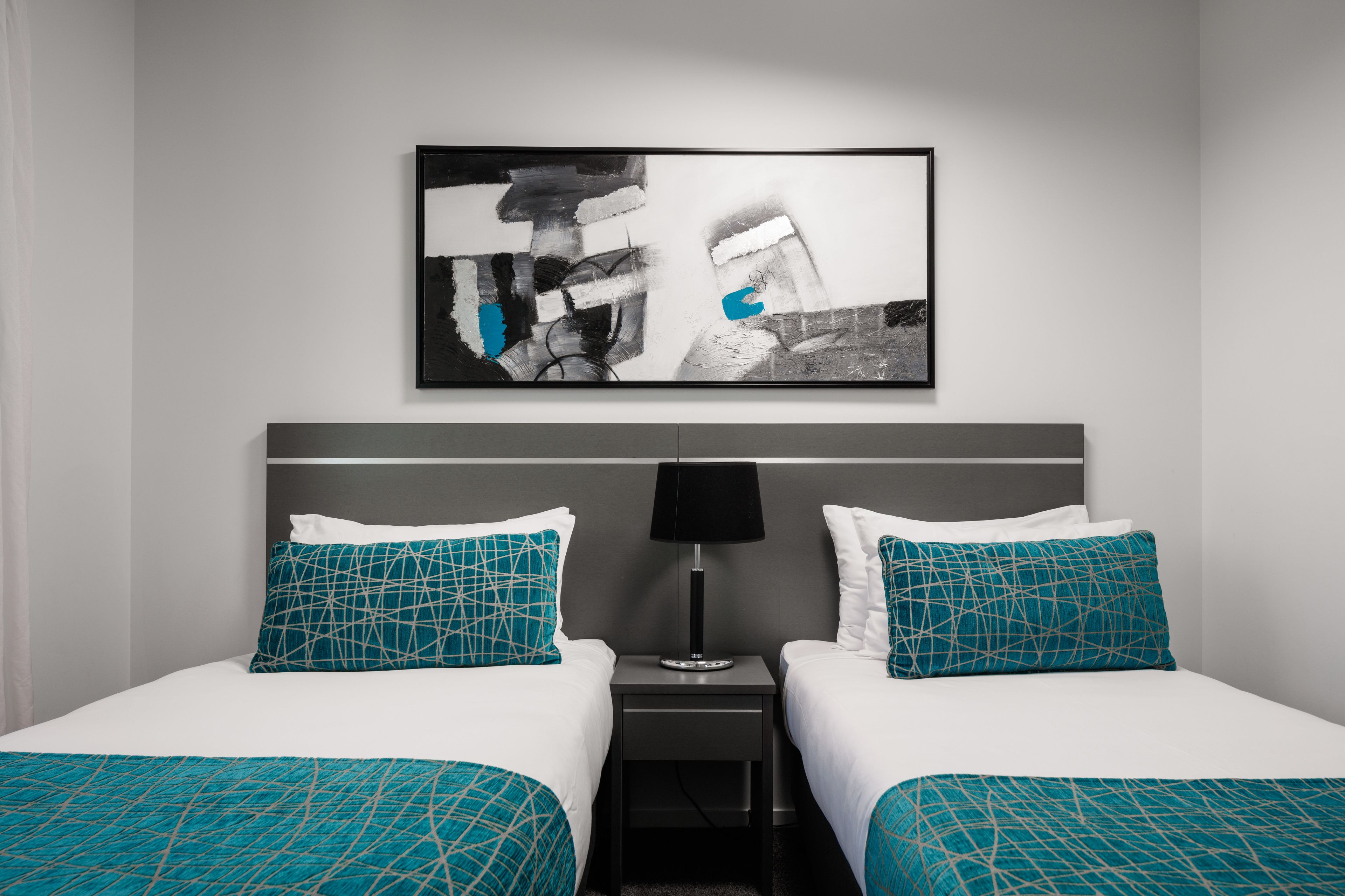 A hotel room with two beds, a nightstand with a lamp, and a large abstract painting on the wall.