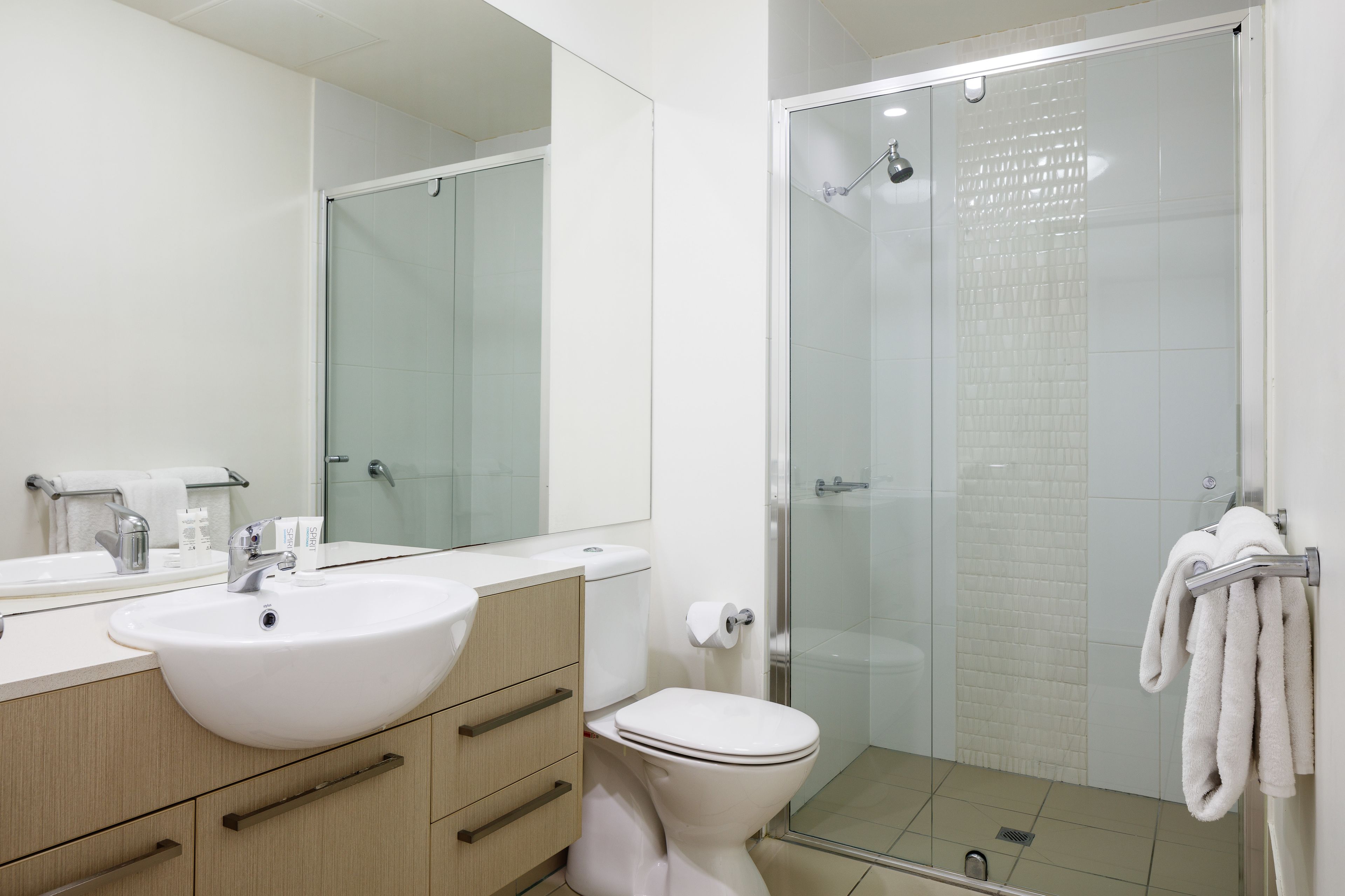 A modern bathroom with a white sink, mirror, and toilet. It includes a glass shower enclosure and a towel rack with towels.