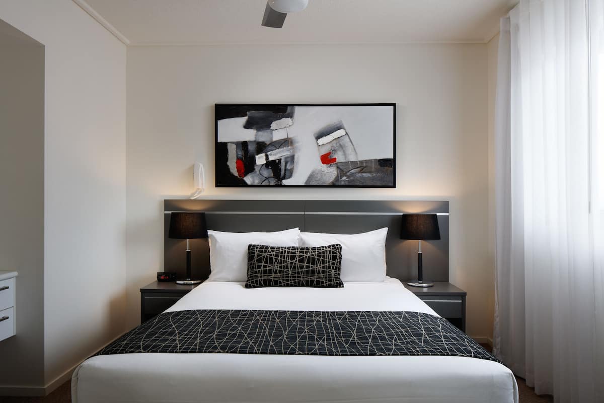 Une chambre d’hôtel comprenant un lit, deux tables de chevet, une œuvre d’art accrochée au mur et une fenêtre avec des rideaux.