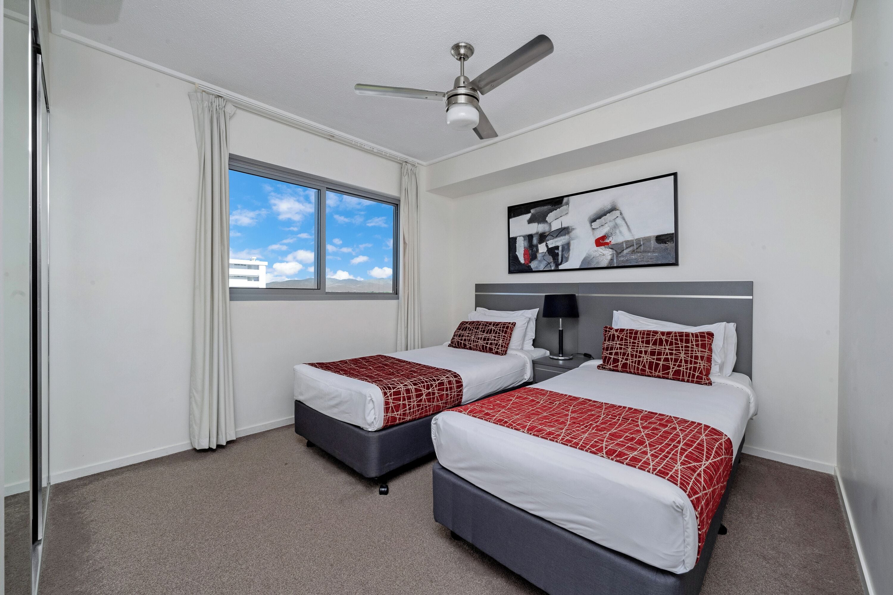 A hotel room with two beds, a ceiling fan, a window with curtains, and a painting on the wall.