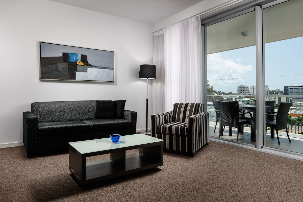Un salon moderne avec un canapé, un fauteuil et une table basse. Un tableau est accroché au mur, et de grandes fenêtres offrent une vue sur la ville.