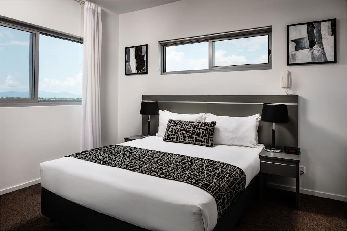 Une chambre d’hôtel bien aménagée, avec un grand lit, deux tables de chevet, une commode, une fenêtre avec des rideaux et deux œuvres d’art encadrées accrochées au mur.