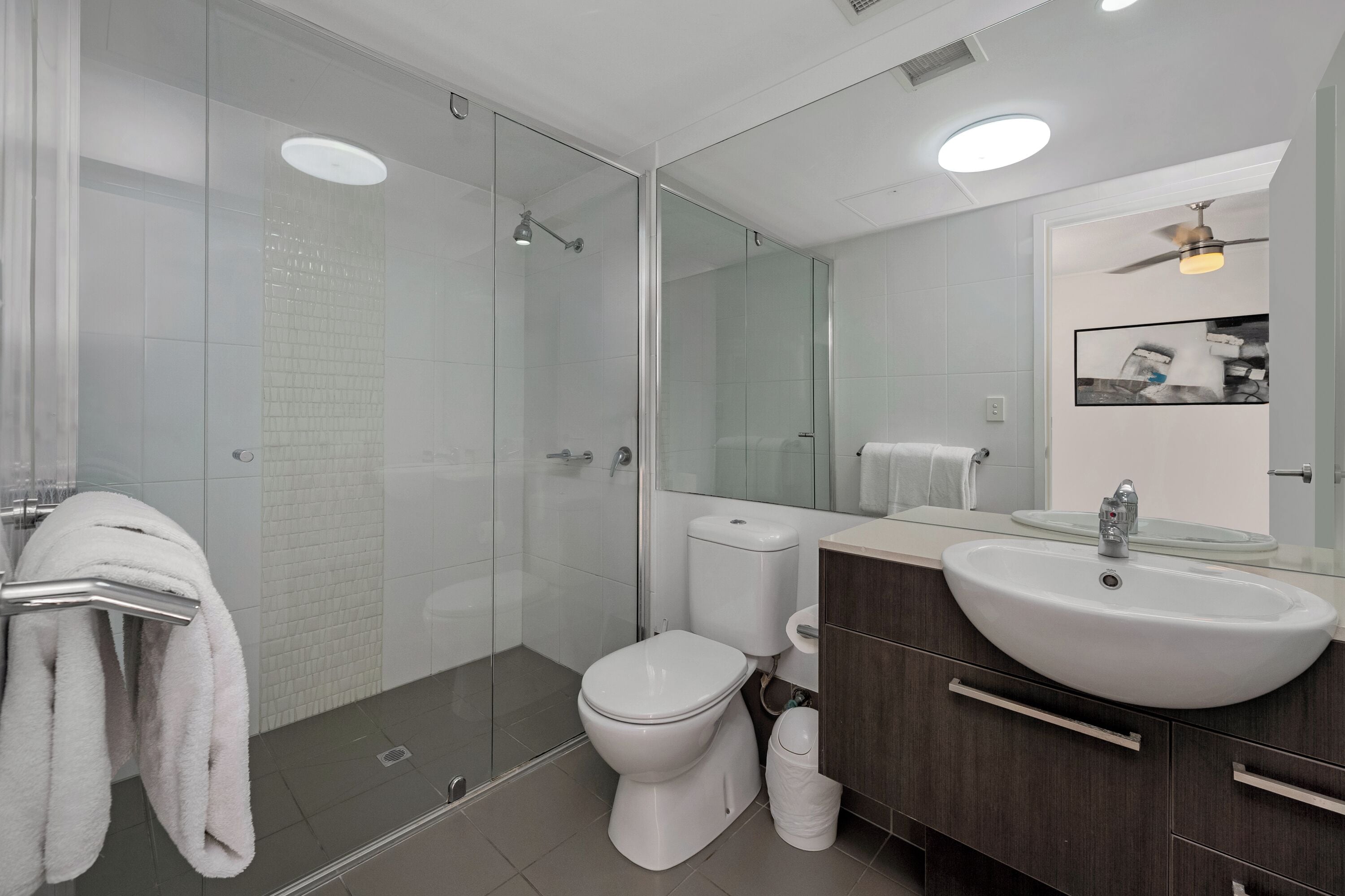 A modern bathroom with a glass-enclosed shower, a white toilet, a sink with a mirror above it, and a ceiling fan.