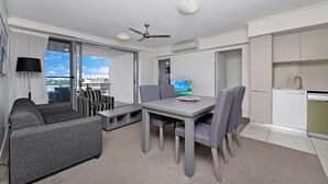 Un salon moderne avec un canapé gris, une table à manger et des chaises, un ventilateur de plafond et une vue sur la ville grâce à de grandes fenêtres.
