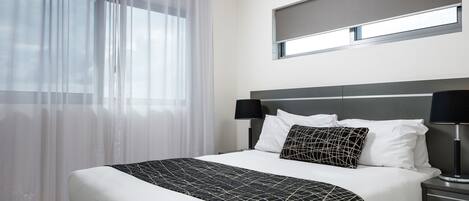 Una cama bien tendida con una colcha de cuadros blancos y negros, dos lámparas negras a cada lado y una ventana con cortinas de encaje.