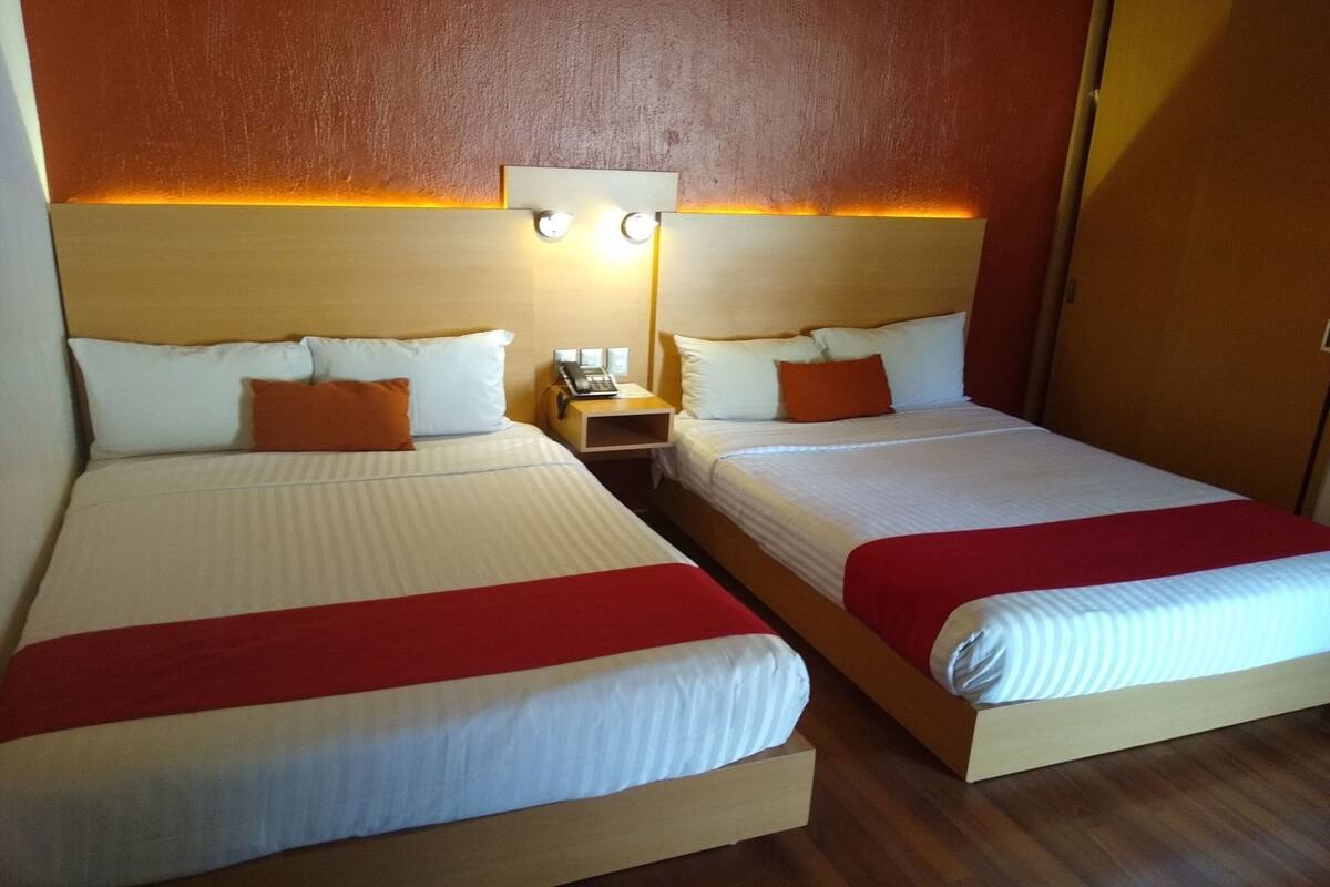 Two double beds with white and red bedding, a nightstand with a phone, and a wall-mounted light fixture.