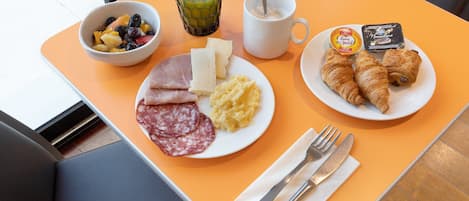 A breakfast tray with croissants, ham, cheese, scrambled eggs, fruit, and juice on an orange table.