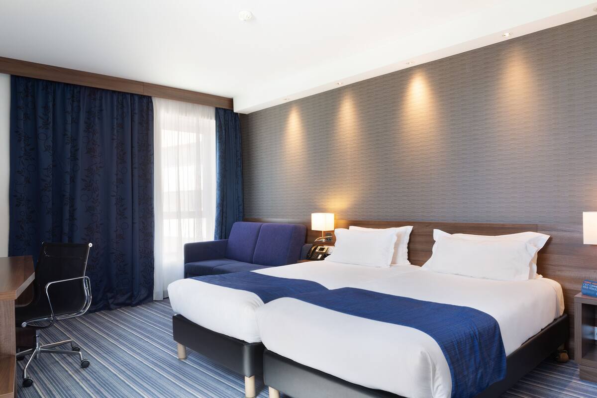 A hotel room with a large bed, a desk, a chair, and a nightstand with a lamp.
