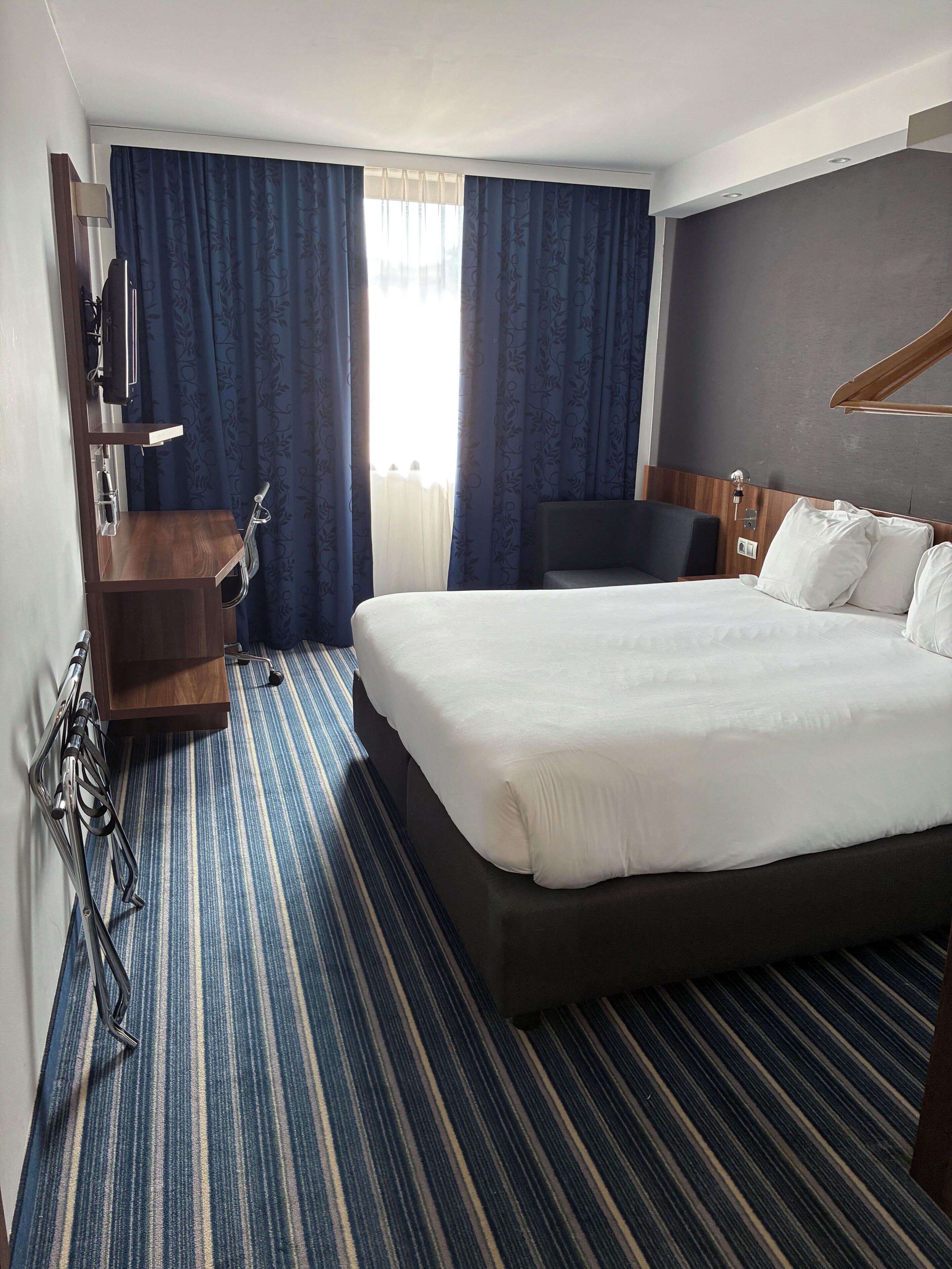 A hotel room with a large bed, a desk with a chair, a TV, and a window with curtains.