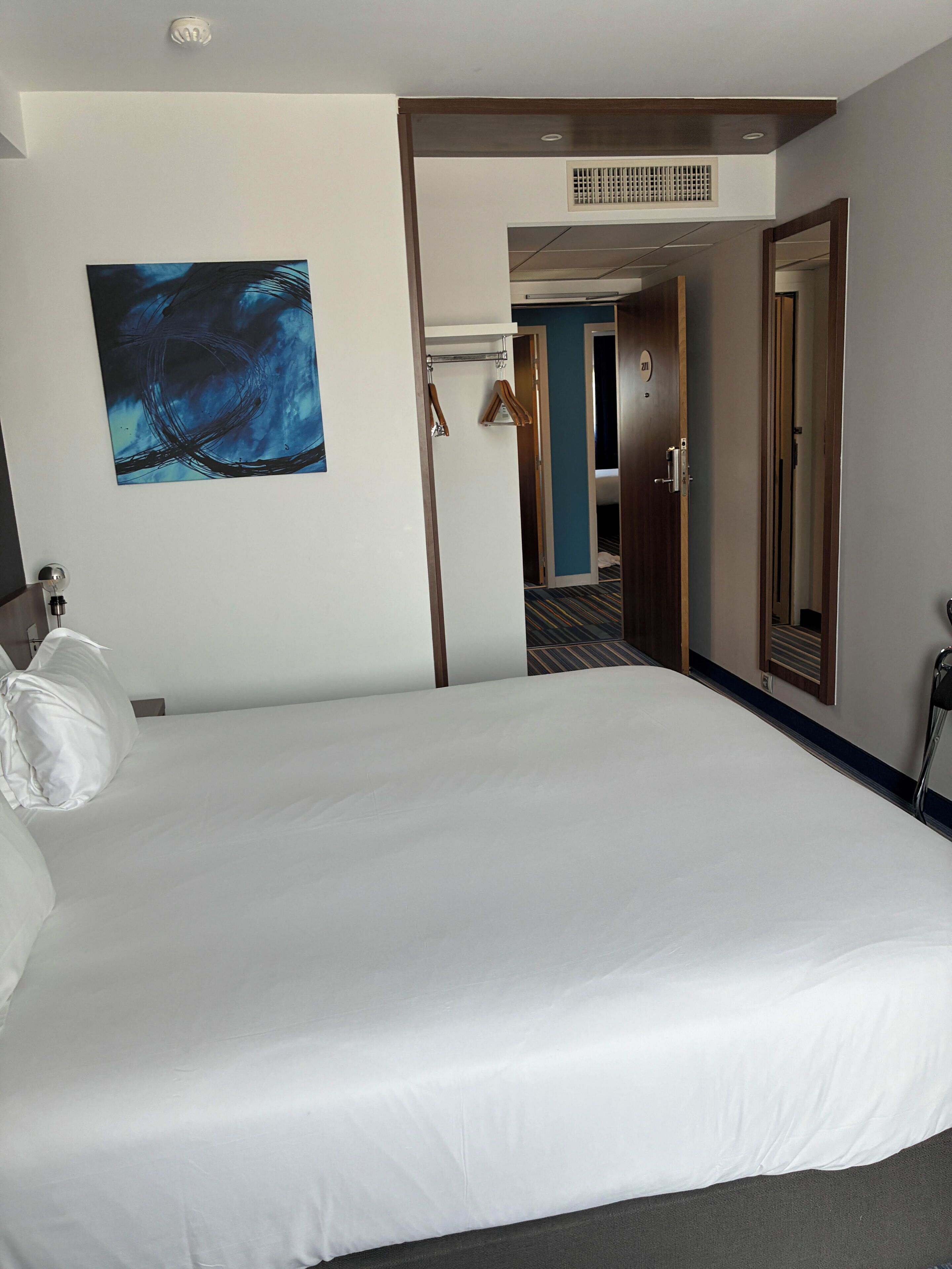A hotel room with a large bed, a painting on the wall, a door leading to another room, and a mirror.