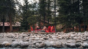 A riverside cabin with a deck, surrounded by large rocks and dense trees.