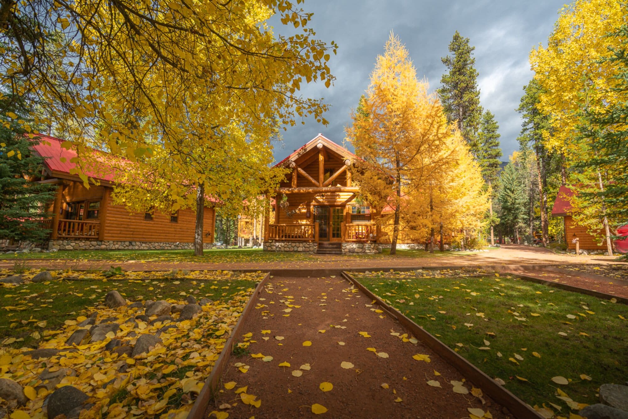A rustic log cabin with a porch, surrounded by autumn foliage and a dirt path.
