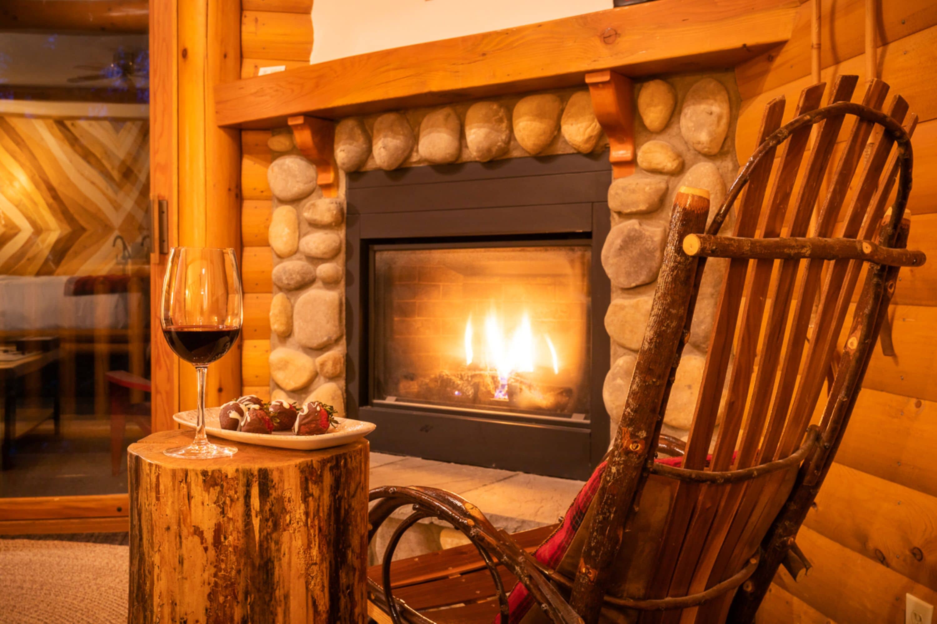A cozy fireplace with a stone surround, a wooden chair, and a plate with food and a glass of red wine on a wooden stump.