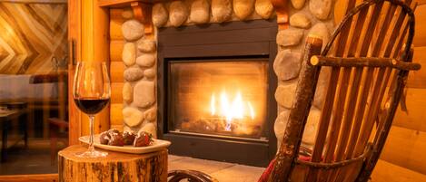 A cozy fireplace with a stone surround, a wooden chair, and a plate with food and a glass of red wine on a wooden stump.