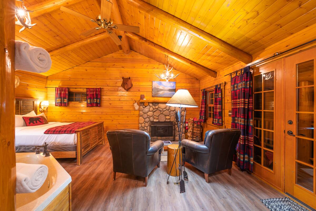 Une chambre aménagée dans un chalet en rondins, avec un lit, deux fauteuils en cuir, une cheminée et un mur en pierre.