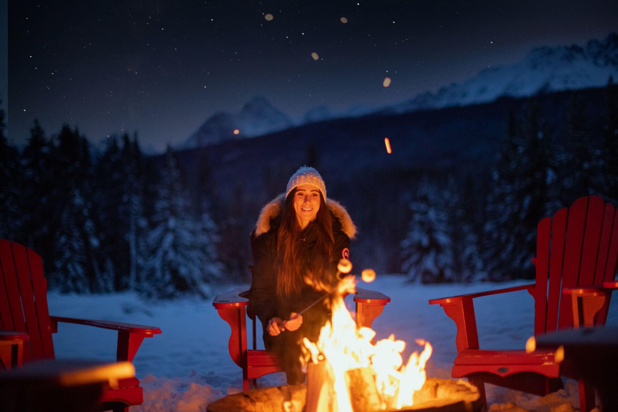 Une personne assise près d’un feu de camp, avec des chaises Adirondack rouges, entourée d’arbres et de montagnes enneigés en arrière-plan.