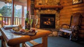 A wooden table with a bottle, glasses, and a cheese board in a cabin with a stone fireplace and a rocking chair.
