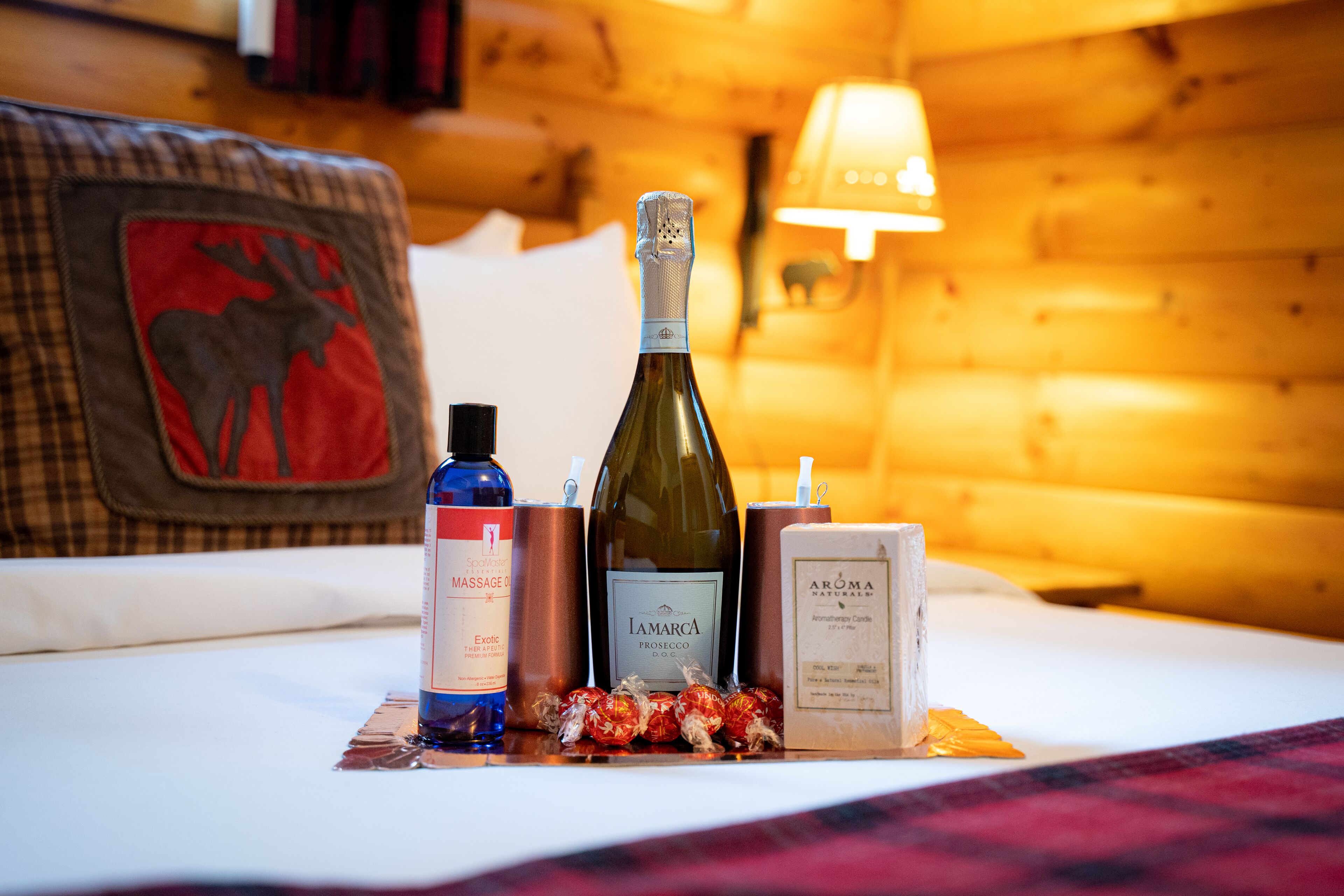 A bed with a red moose patch, a bottle of wine, a massage oil bottle, and a candle on a plaid blanket.