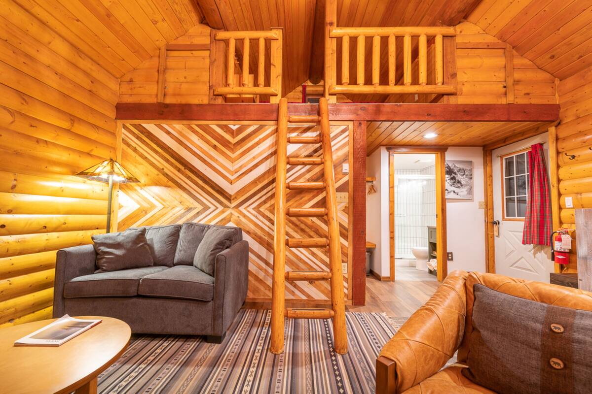 L’intérieur d’une cabane en rondins, avec un escalier en bois, deux canapés, un tapis à motifs et une table en bois.