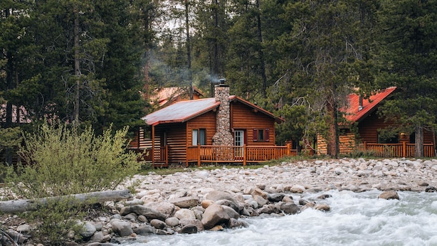 A log cabin with a stone chimney, surrounded by a rocky riverbank and dense forest.