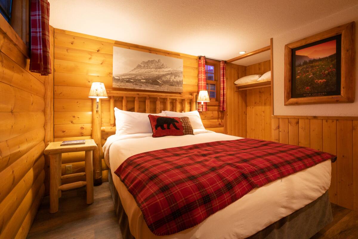Une pièce aménagée comme un chalet en bois, avec un lit, deux lampes de chevet, un petit tabouret en bois et un tableau encadré au mur.