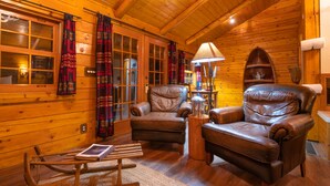 A cozy log cabin interior with two leather armchairs, a wooden rocking chair, and a lamp.