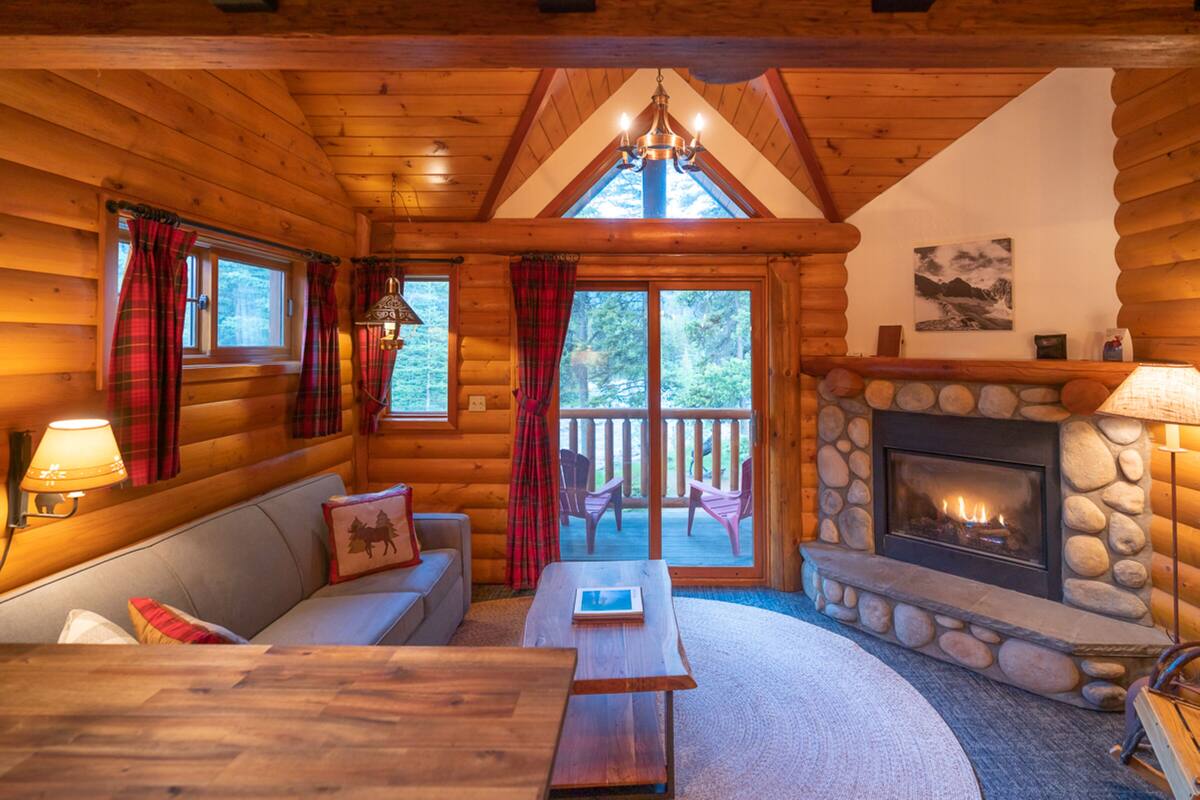 L’intérieur d’une cabane en rondins, avec une cheminée en pierre, un plafond en bois et un coin salon confortable comprenant un canapé et une table basse.