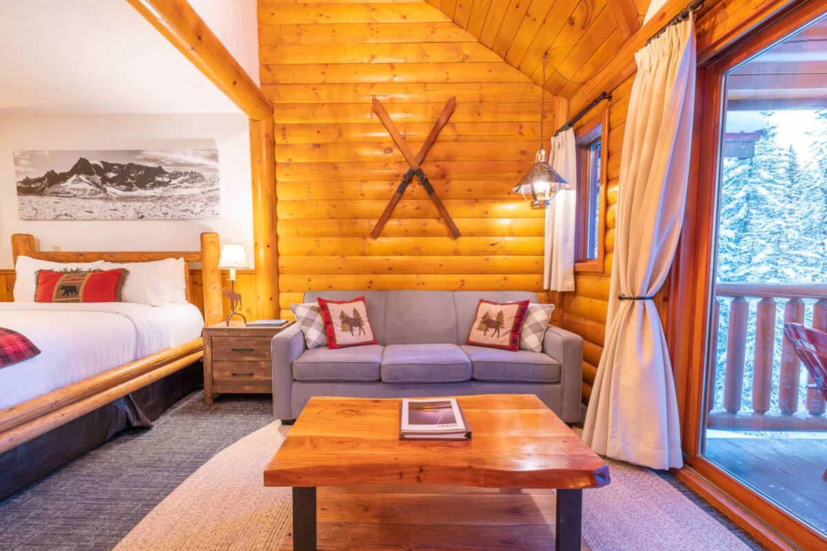 Une pièce chaleureuse aménagée dans un chalet en bois, avec un lit, un canapé, une table basse en bois et une grande fenêtre avec des rideaux.