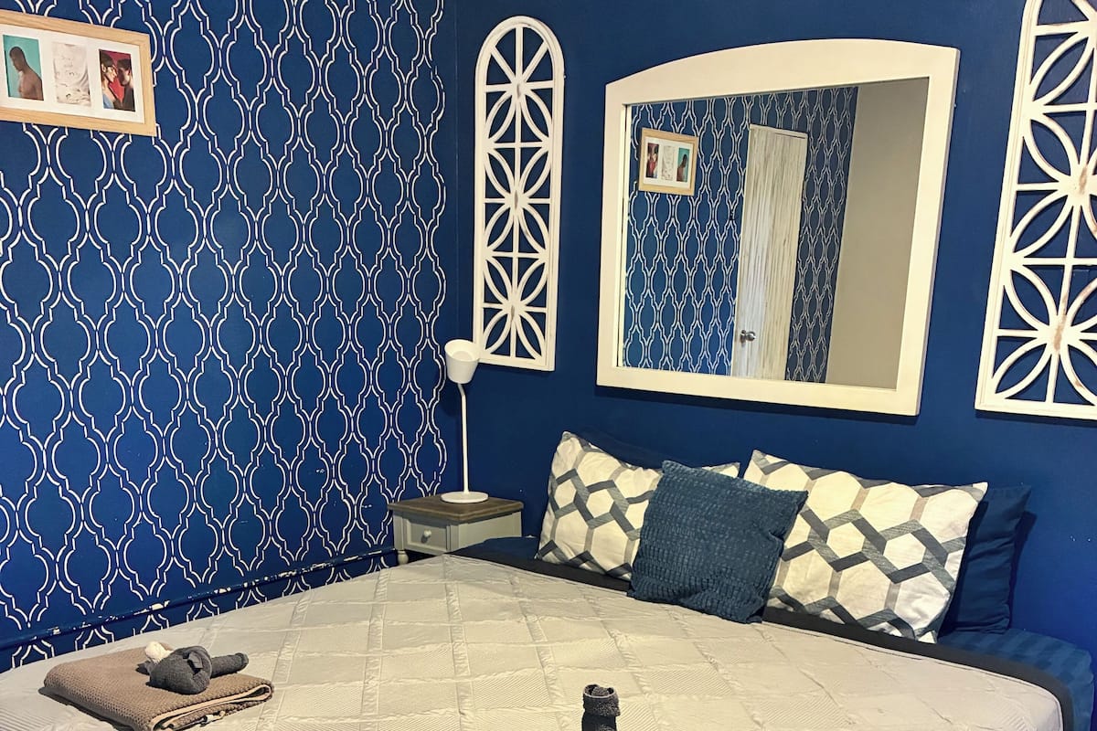 A bedroom with a bed, a nightstand, a mirror, and patterned wallpaper.