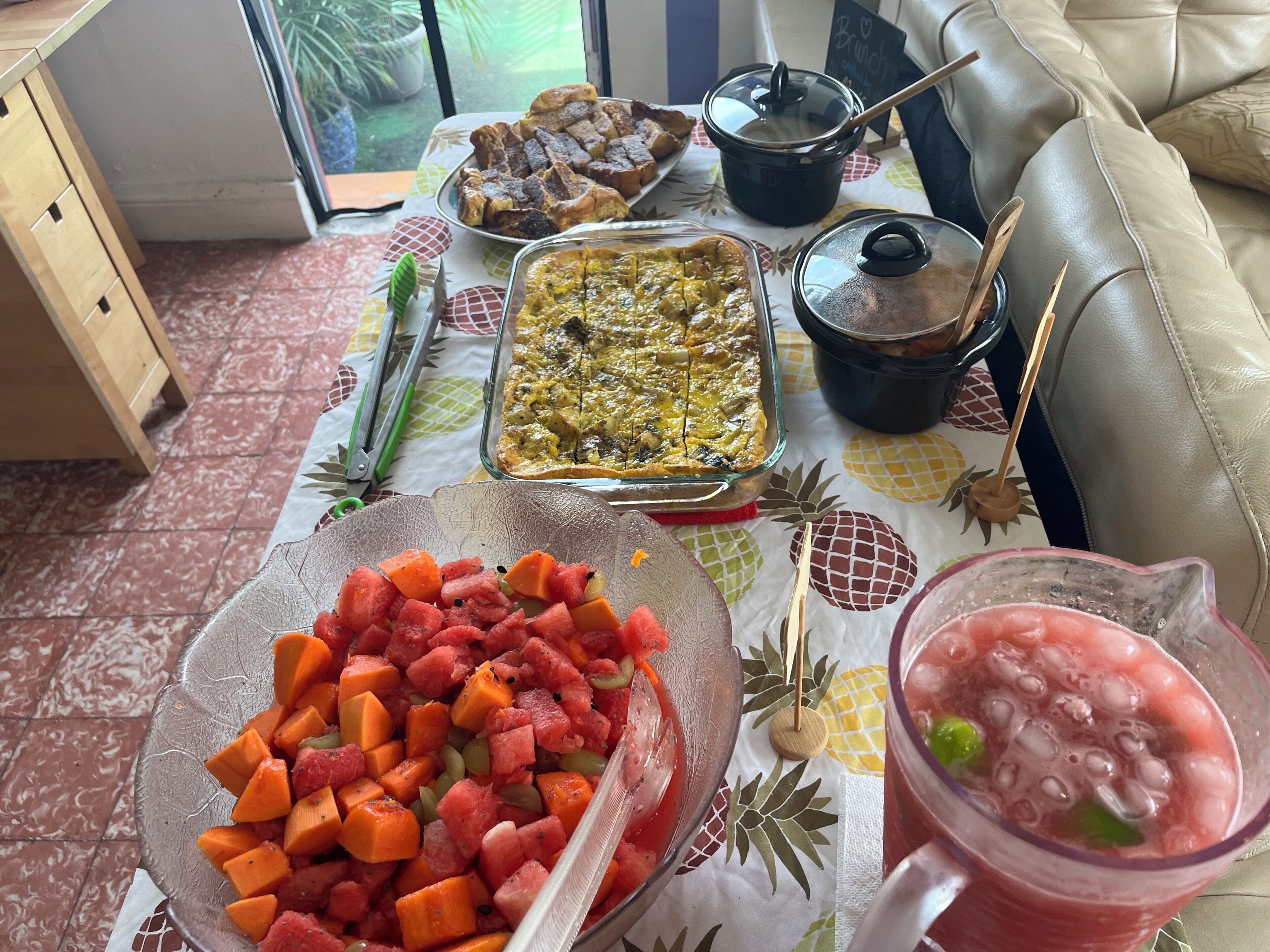 A dining table with various dishes, including a fruit salad, a casserole, and a drink, set on a patterned tablecloth.