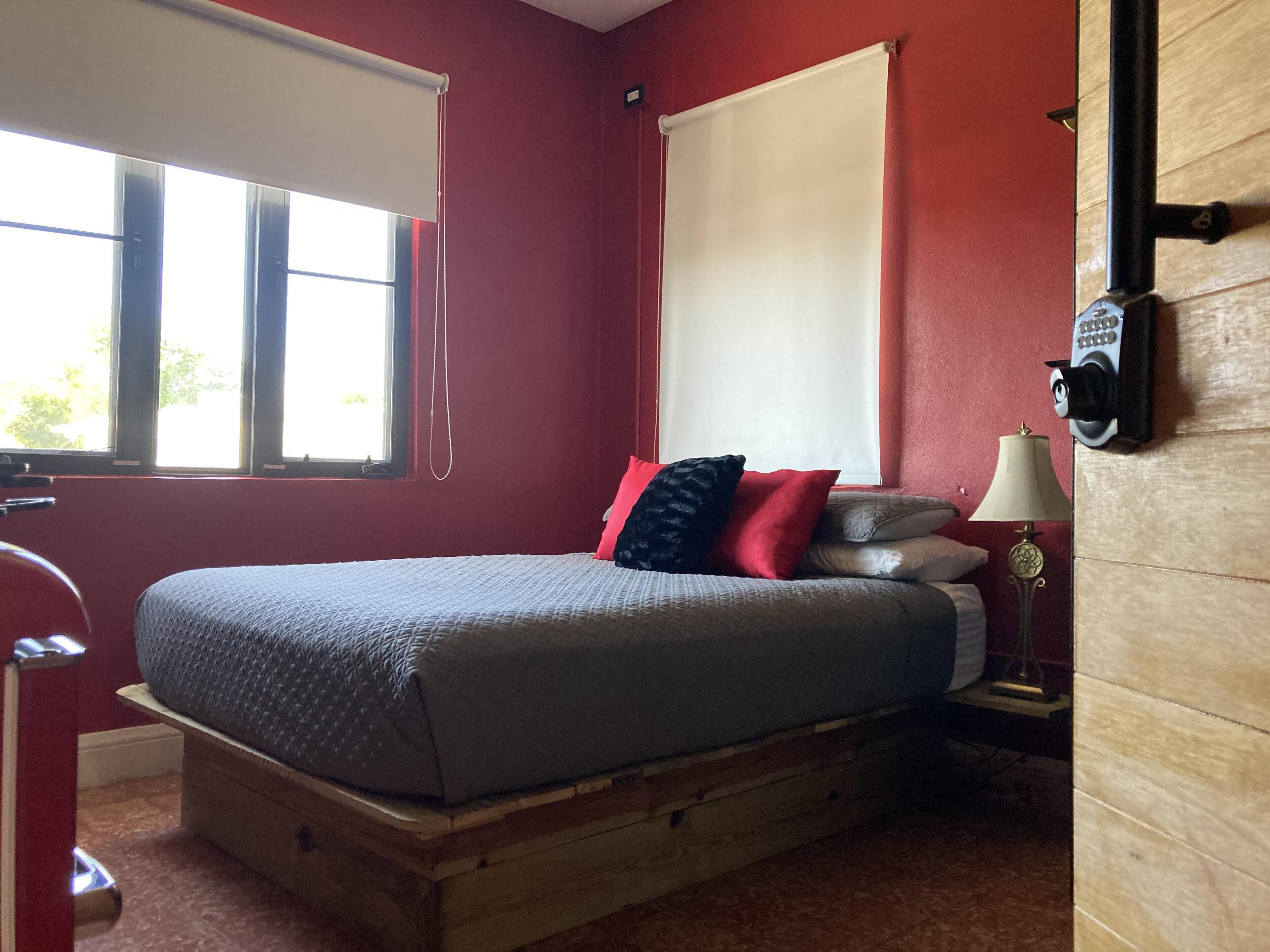 A bedroom with a wooden bed, red walls, a window with blinds, a bedside lamp, and a wall-mounted thermostat.