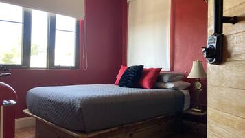 A bedroom with a wooden bed, red walls, a window with blinds, a bedside lamp, and a wall-mounted thermostat.