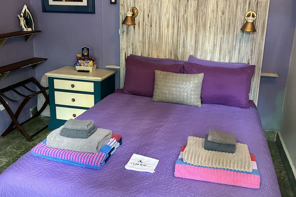 A bedroom with a purple bedspread, two pillows, a wooden headboard, a nightstand with a lamp, and a framed painting on the wall.