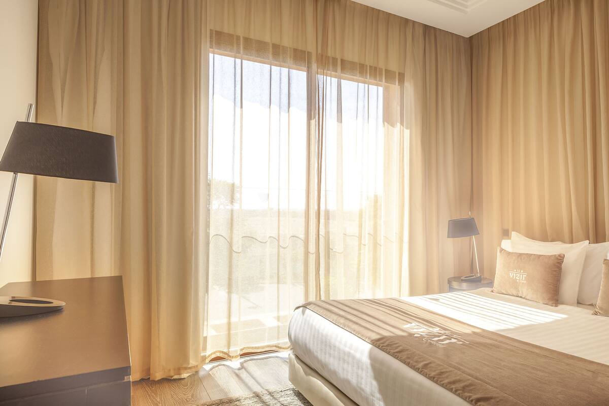 A hotel room with a bed, a desk, a lamp, and a window with curtains.