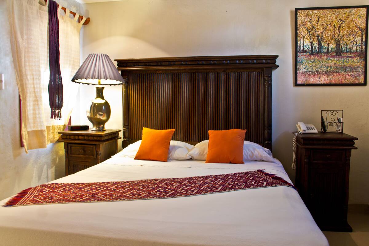 A hotel room with a large bed, two orange pillows, a red and white patterned throw, a lamp on a bedside table, a telephone, and a painting on the wall.