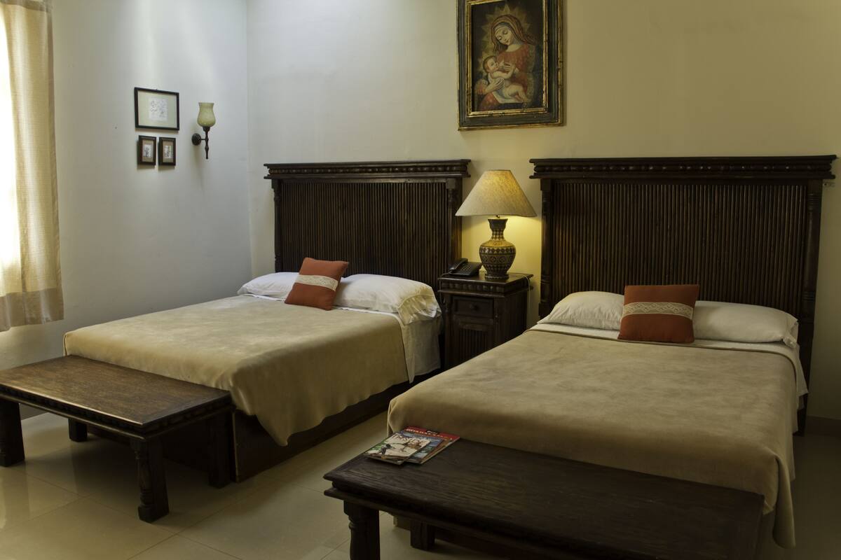 A hotel room with two single beds, a nightstand with a lamp, a bench, and a framed picture on the wall.